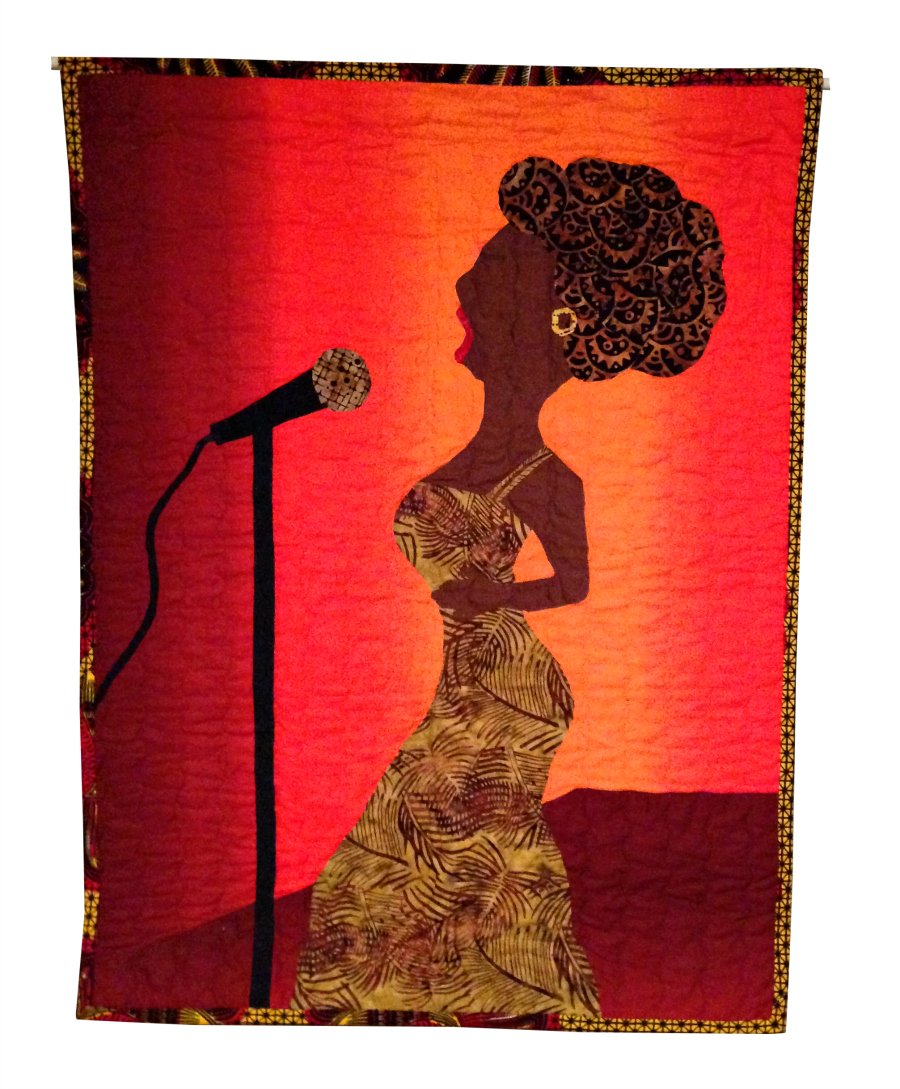 Wambui Made It:'Singing Solo' quilt (front) -- If you have ever had to face the music alone. -- This quilt is a decorative wall quilt. I made this quilt in 2012. This quilt began as a hand drawing of a nightclub singer. I then traced that drawing and cut it into pieces like a puzzle. I then used the paper pieces as guides to cut various cotton fabrics into the shapes of the head, the dress, the microphone, etc. I pieced those fabrics together to form the picture - out of fabric. I then sewed the fabric pieces together on the orange gradient fabric.